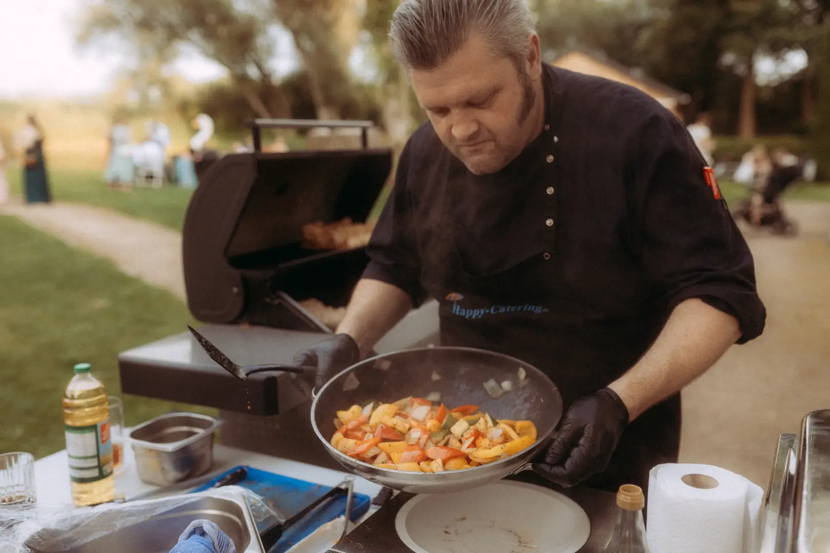 Ein Koch in schwarzer Arbeitskleidung mit „Happy-Catering“-Logo bereitet draußen bei einer Hochzeit frisches Pfannengemüse in einem Wok zu. Im Hintergrund sind Gäste im Grünen zu sehen. Professionelle Begleitung durch einen Hochzeitsfotograf in Schwerin und MV.