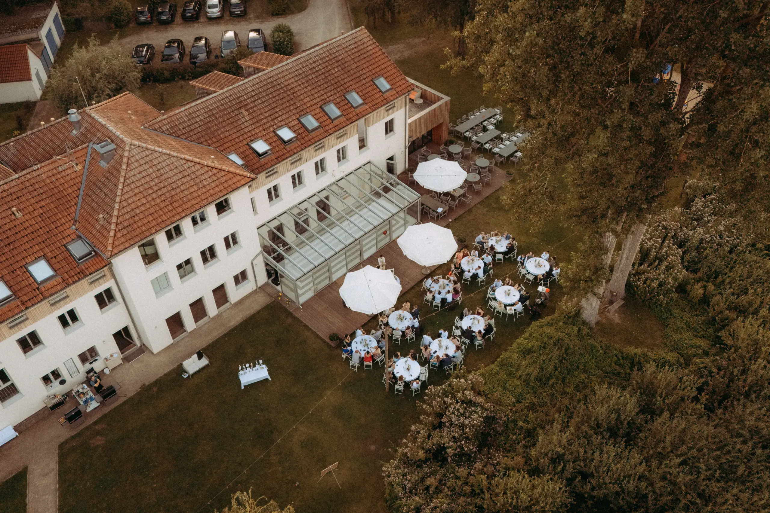 Luftaufnahme einer Hochzeitsfeier im Freien mit weißen runden Tischen und Sonnenschirmen auf einer grünen Wiese neben einem historischen Gebäude. Aufgenommen von einem Hochzeitsfotograf in Schwerin und MV, zeigt das Bild eine elegante Gartenhochzeit in einer malerischen Umgebung. Luftaufnahme einer Hochzeitsfeier im Freien mit weißen runden Tischen und Sonnenschirmen auf einer grünen Wiese neben einem historischen Gebäude. Aufgenommen von einem Hochzeitsfotograf in Schwerin und MV, zeigt das Bild eine elegante Gartenhochzeit in einer malerischen Umgebung.