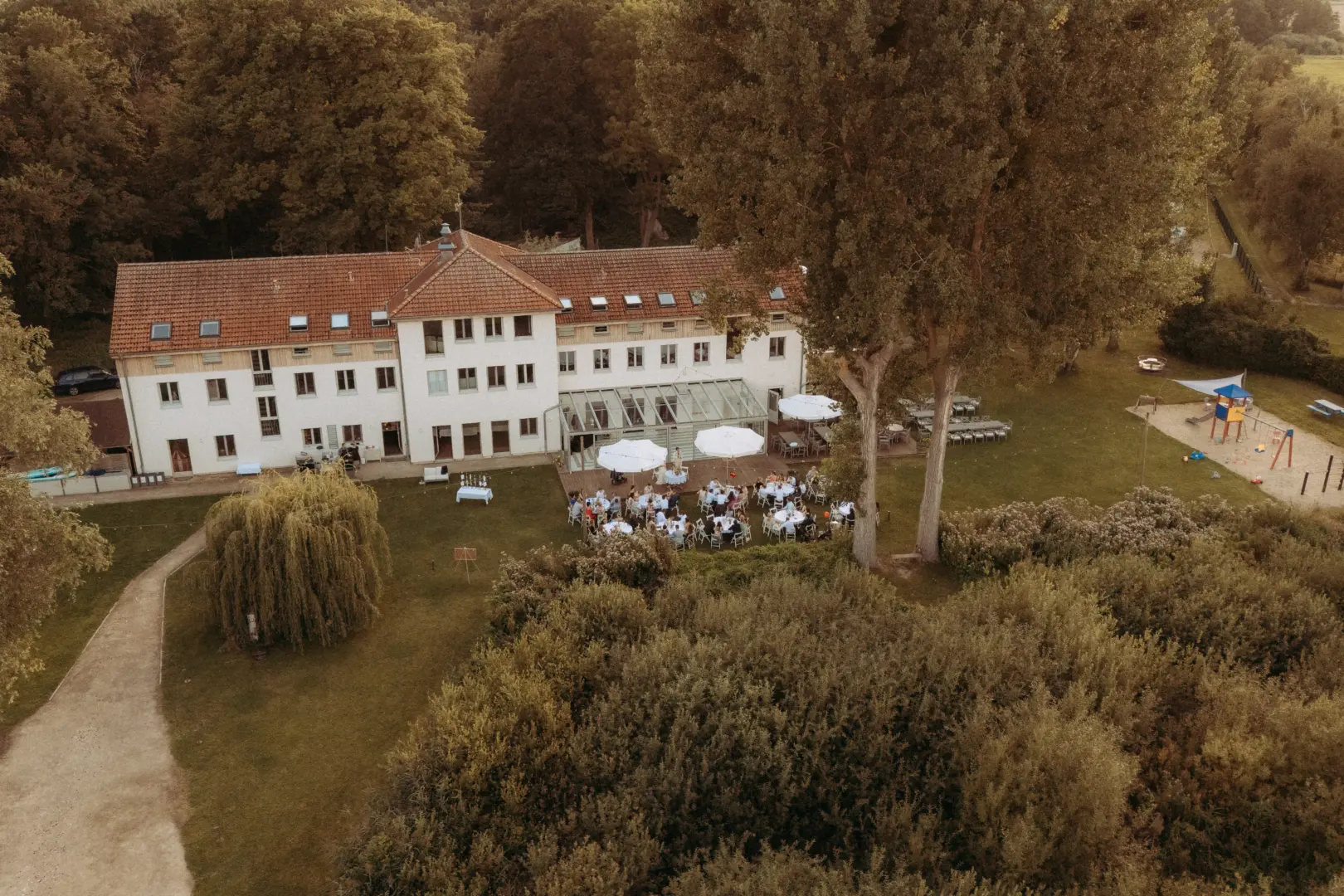 Luftaufnahme einer Hochzeitsfeier im Freien vor einem historischen Herrenhaus mit Ziegeldach. Gäste sitzen an runden Tischen unter weißen Sonnenschirmen auf einer Terrasse, umgeben von weitläufigen Grünflächen und Bäumen. Hochzeitsfotograf in Schwerin und MV.