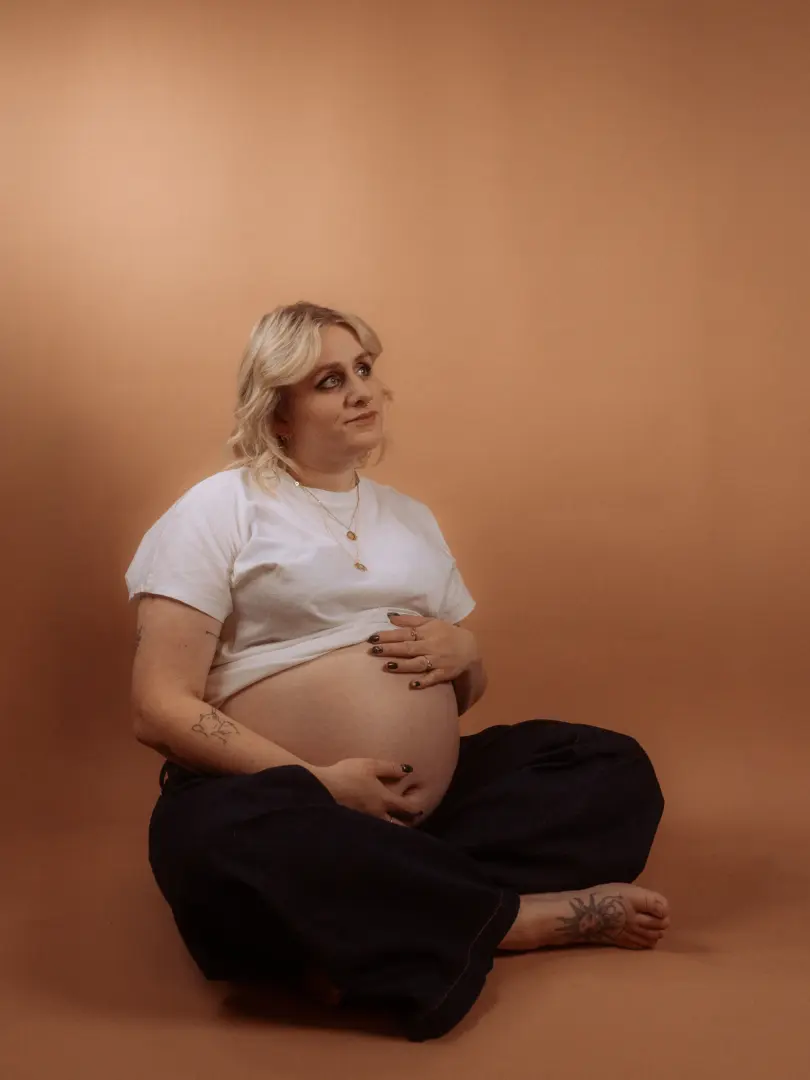 Eine schwangere Frau mit blondem Haar sitzt nachdenklich im Schneidersitz vor einem warmen, beige-farbenen Hintergrund in einem Fotostudio in Schwerin. Sie trägt ein weißes T-Shirt und eine dunkle Hose, ihre Hände ruhen liebevoll auf ihrem Babybauch.