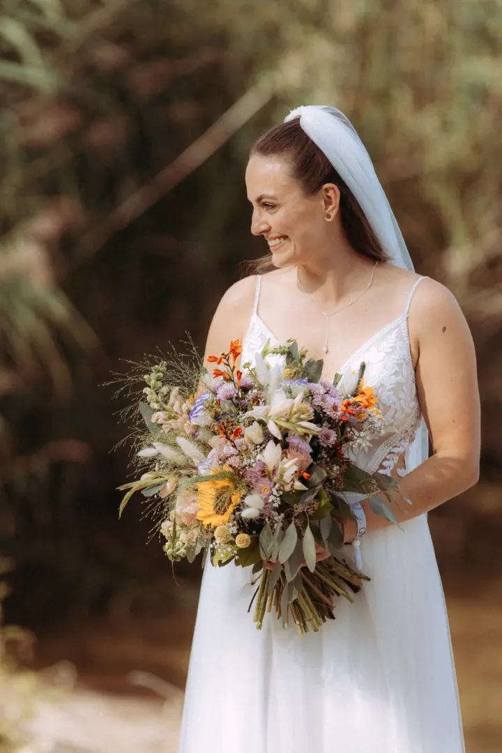 Eine strahlende Braut im weißen Spitzenkleid hält einen farbenfrohen Wildblumenstrauß in den Händen. Sie lächelt glücklich zur Seite, während ihr Schleier sanft im Wind weht. Ein stimmungsvolles Porträt für den Hochzeitsfotograf in Schwerin und MV.