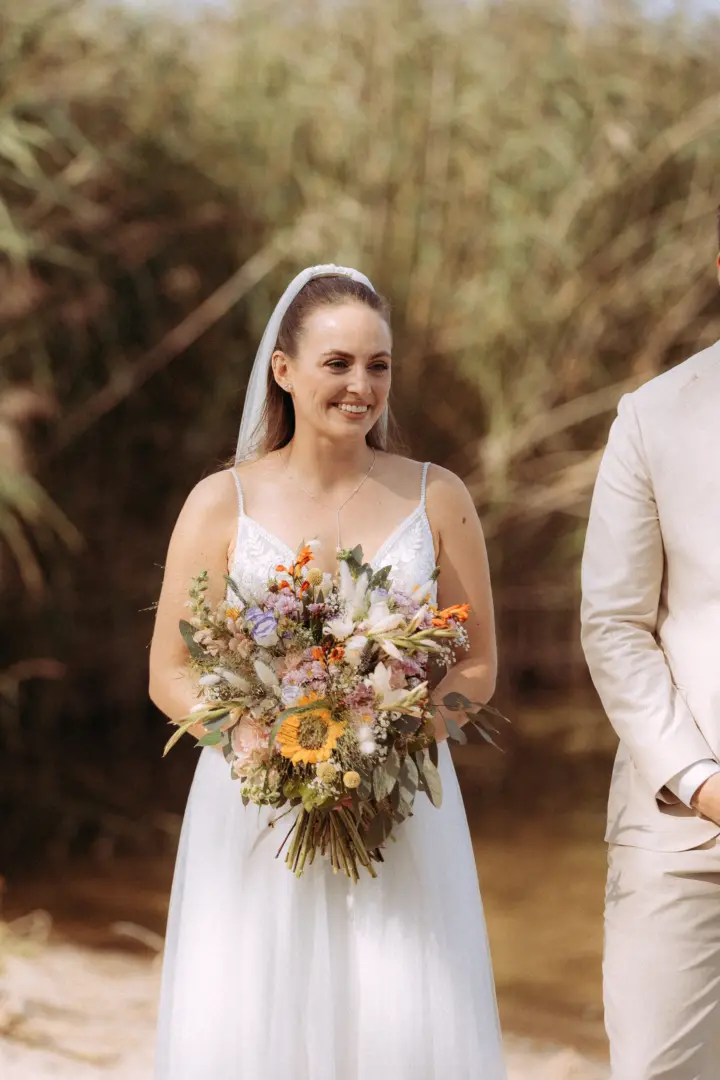 Eine strahlende Braut im weißen Kleid hält bei ihrer Hochzeitszeremonie einen großen, farbenfrohen Wildblumenstrauß mit Sonnenblumen. Im Hintergrund ist ihr Bräutigam in einem beigen Anzug zu sehen. Professionelle Aufnahmen vom Hochzeitsfotograf in Schwerin und MV.