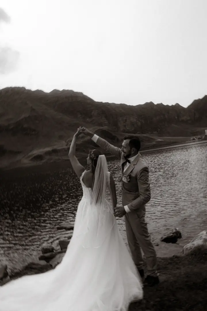 Ein Brautpaar tanzt romantisch vor einer malerischen Bergkulisse an einem See. Die Braut trägt ein elegantes Hochzeitskleid mit Schleier, während der Bräutigam im Anzug führt. Kinematischer Hochzeitsfotograf in der Schweiz.
