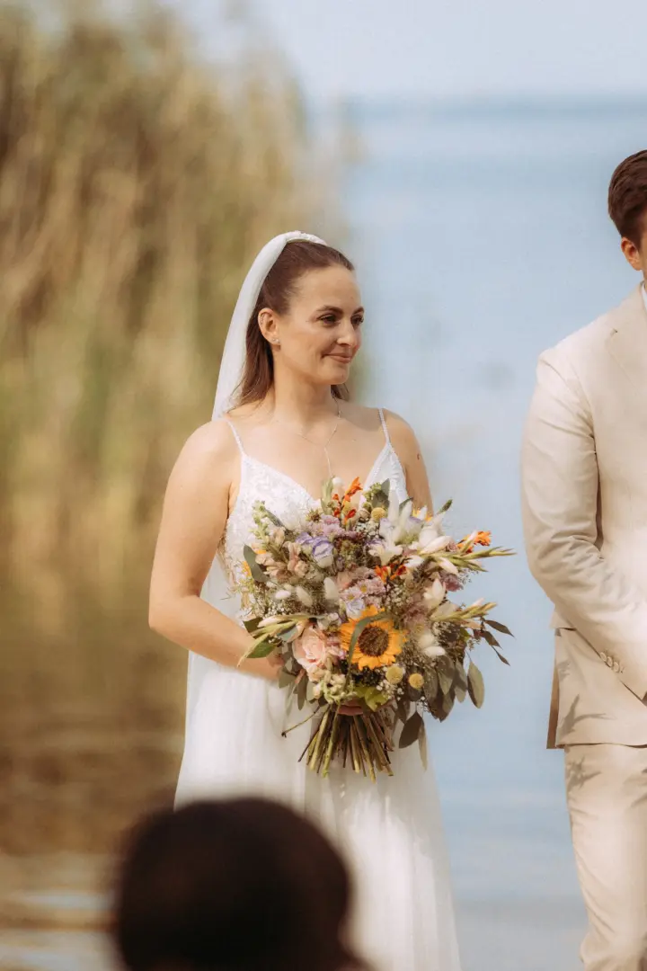 Eine glückliche Braut in einem weißen Brautkleid hält einen großen, bunten Wildblumenstrauß mit Sonnenblumen in den Händen, während sie neben ihrem Bräutigam an einem See steht. Ein stimmungsvoller Moment festgehalten von einem Hochzeitsfotograf in Schwerin und MV.