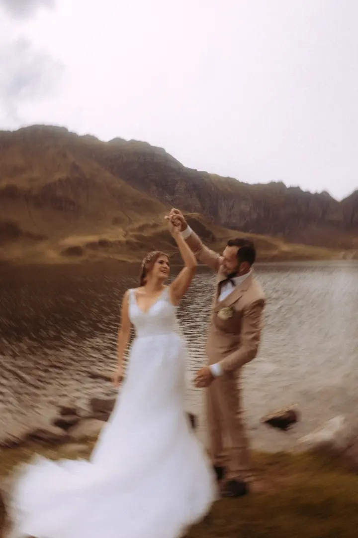 Ein Brautpaar tanzt glücklich am Ufer eines Bergsees vor einer dramatischen Gebirgskulisse. Die Braut trägt ein weißes Hochzeitskleid, der Bräutigam einen beigen Anzug. Kinematischer Hochzeitsfotograf in der Schweiz, der diesen romantischen Moment in einer malerischen Landschaft festhält.