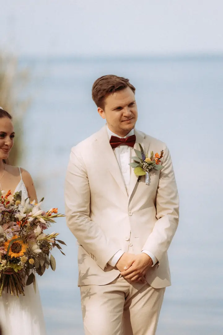 Bräutigam in einem hellen beigen Anzug mit weinroter Fliege und floralem Anstecker steht während der Zeremonie neben der Braut mit ihrem bunten Blumenstrauß. Ein emotionaler Moment festgehalten von einem Hochzeitsfotograf in Schwerin und MV vor einer ruhigen Wasserlandschaft.