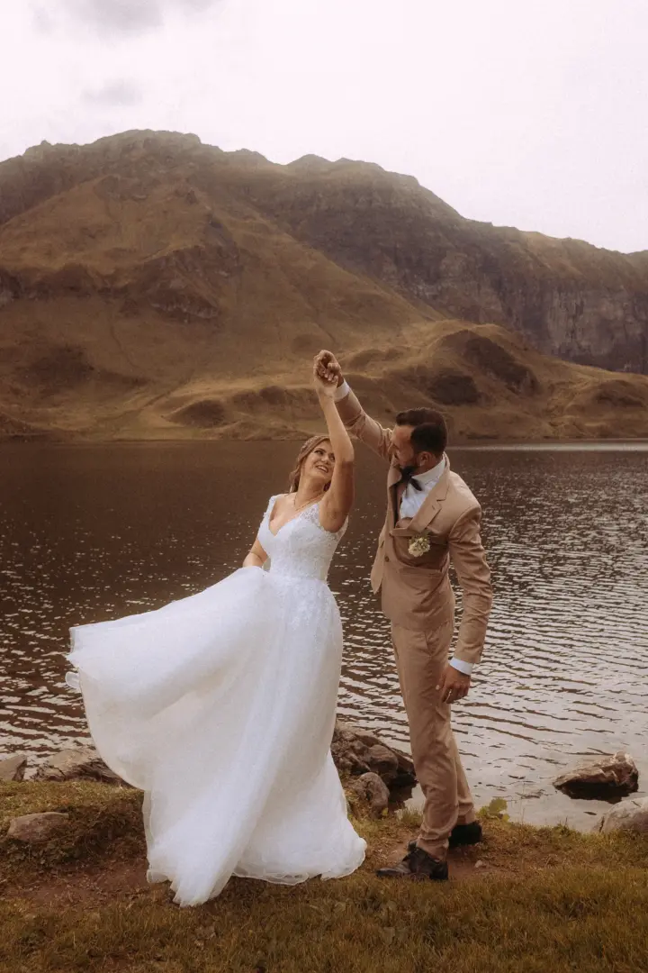 Ein Brautpaar tanzt glücklich am Ufer eines Bergsees vor einer imposanten Bergkulisse. Die Braut trägt ein weißes Spitzenkleid, während der Bräutigam einen beigen Anzug trägt. Ein kinematischer Hochzeitsfotograf in der Schweiz hat diesen romantischen Moment festgehalten.