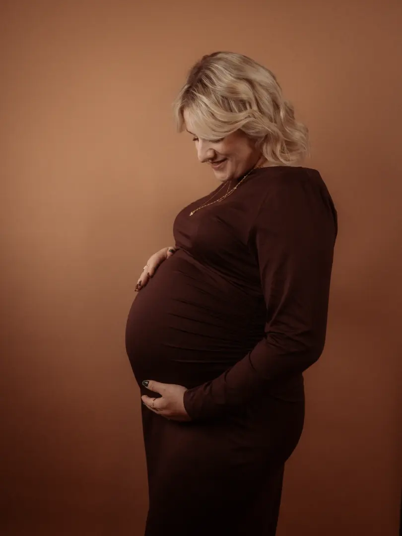 Eine schwangere Frau mit blondem Haar trägt ein elegantes, dunkelbraunes Kleid und hält liebevoll ihren Babybauch. Die Aufnahme entstand in einem Fotostudio in Schwerin vor einem warmen, braunen Hintergrund und strahlt eine ruhige, emotionale Atmosphäre aus.