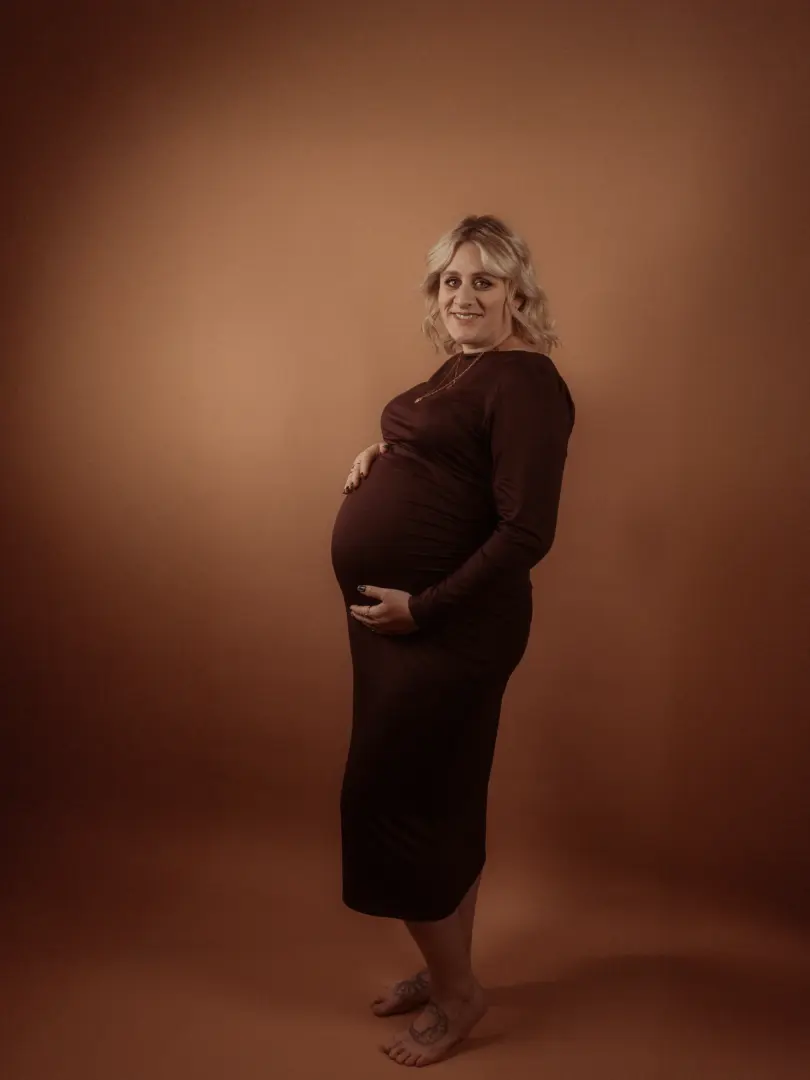 Eine schwangere Frau mit blonden Haaren trägt ein elegantes, dunkelbraunes Kleid und lächelt sanft in die Kamera, während sie liebevoll ihren Babybauch hält. Die Aufnahme entstand in einem professionellen Fotostudio in Schwerin vor einem warmen, braunen Hintergrund.