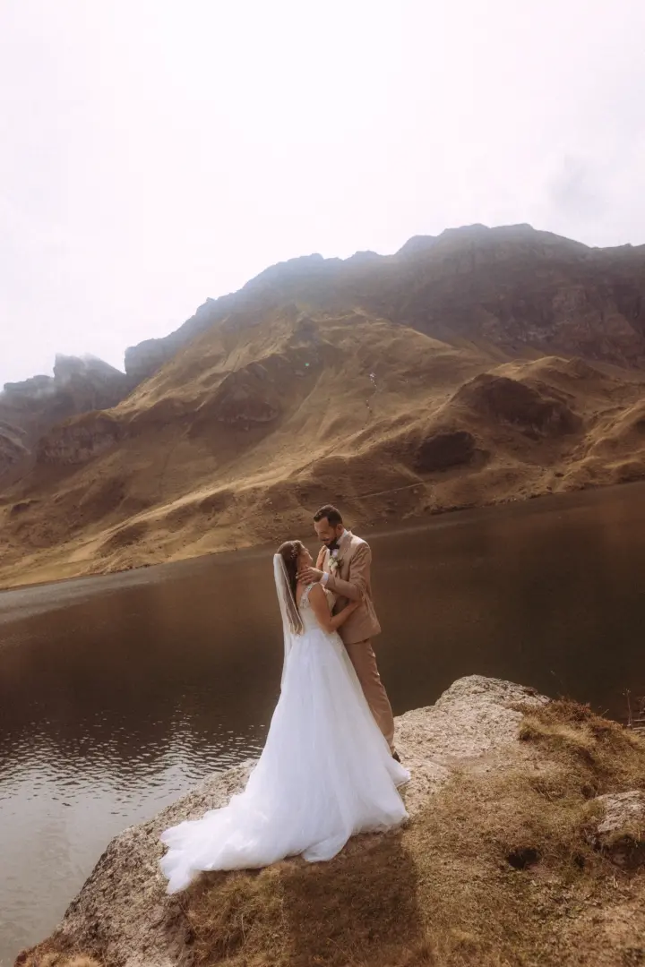 Ein Brautpaar in Hochzeitskleidung steht sich liebevoll gegenüber auf einem Felsvorsprung vor einem malerischen Bergsee und imposanten Bergen. Kinematischer Hochzeitsfotograf in der Schweiz.