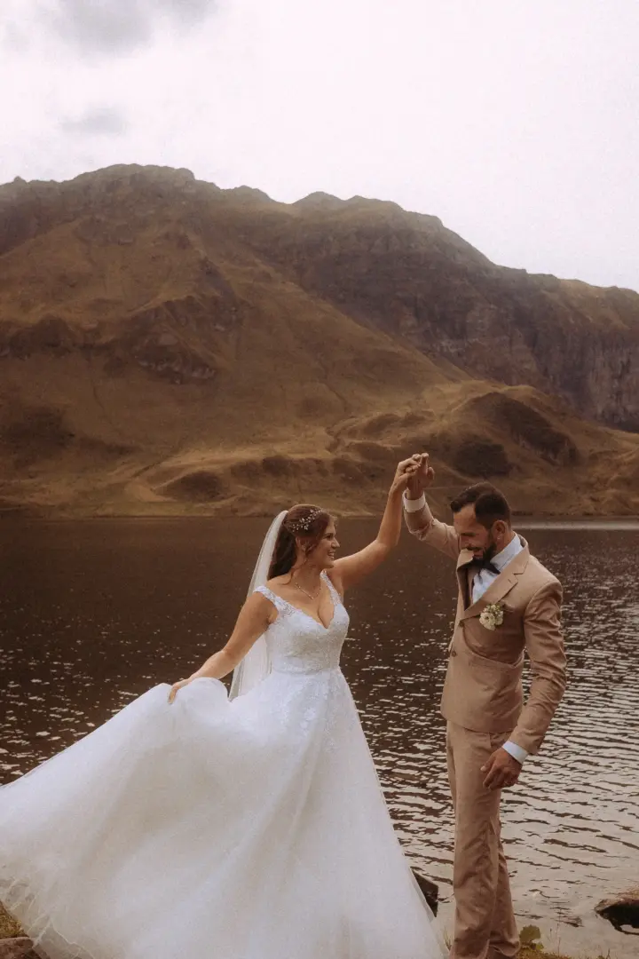 Ein glückliches Brautpaar tanzt an einem See vor einer imposanten Bergkulisse. Die Braut trägt ein weißes Spitzenkleid mit Schleier, der Bräutigam einen beigen Anzug. Ein kinematischer Hochzeitsfotograf in der Schweiz hat diesen romantischen Moment festgehalten.