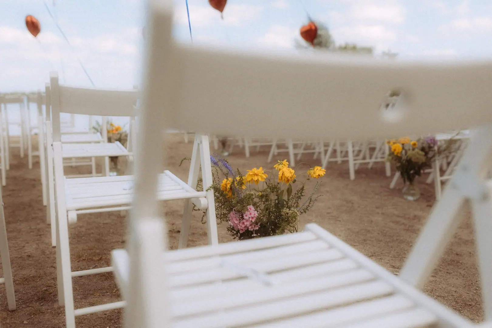 Weiße Klappstühle für eine Hochzeitszeremonie im Freien am See, dekoriert mit bunten Sommerblumen in Vasen. Die romantische Kulisse unter freiem Himmel bietet eine inspirierende Szene für Paare, die einen Hochzeitsfotograf in Schwerin und MV suchen.