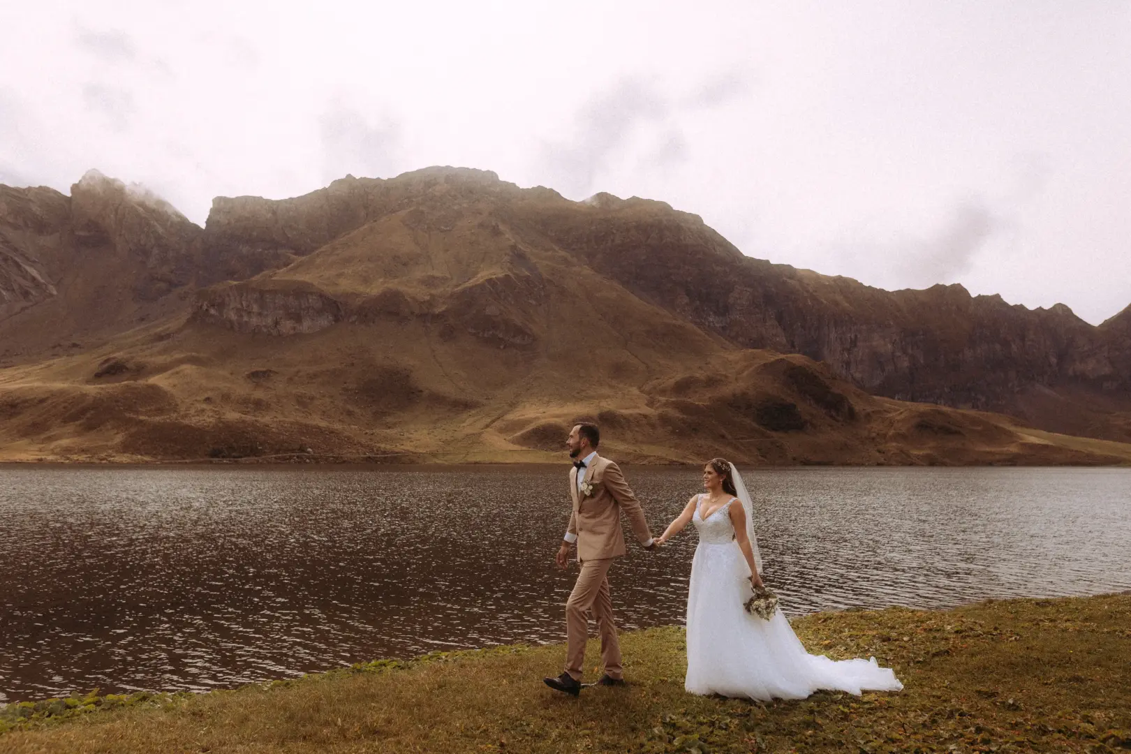 Ein Brautpaar hält Händchen und spaziert vor einer dramatischen Bergkulisse an einem Seeufer entlang. Die Braut trägt ein weißes Hochzeitskleid, der Bräutigam einen beigen Anzug. Kinematischer Hochzeitsfotograf in der Schweiz für unvergessliche Hochzeitsmomente in der Natur.