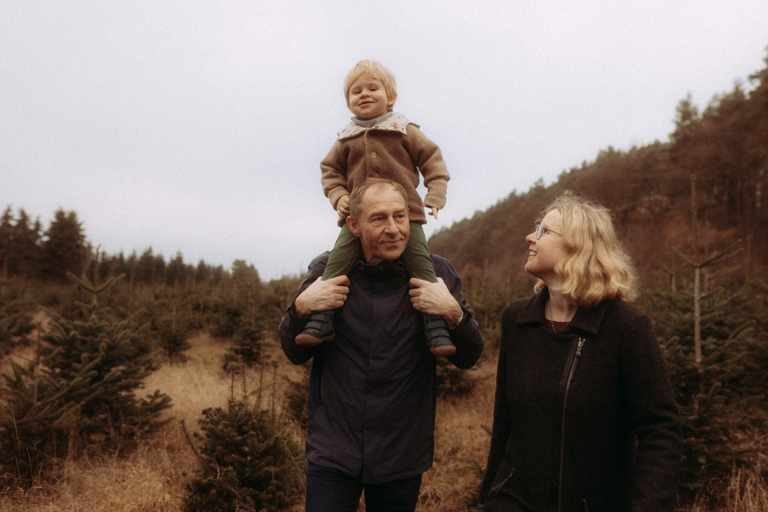 Eine Familie spaziert durch eine winterliche Tannenbaumschonung, das kleine Kind sitzt beim Vater auf den Schultern.