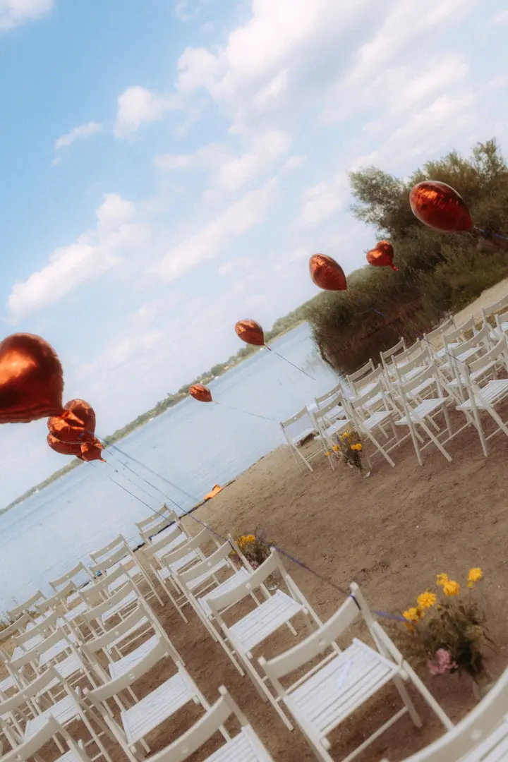 Eine romantische Hochzeitszeremonie am See mit weißen Stühlen, die im Sand aufgereiht sind, geschmückt mit glänzenden, herzförmigen roten Luftballons und Blumenarrangements, fotografiert von einem Hochzeitsfotograf Müritz und MV.