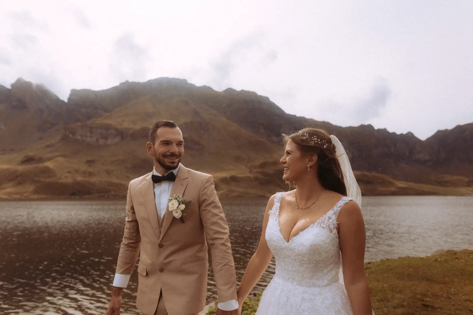 Ein glückliches Brautpaar hält Händchen vor einer dramatischen Bergkulisse an einem See. Der Bräutigam trägt einen beigen Anzug, die Braut ein weißes Spitzenkleid. Kinematischer Hochzeitsfotograf in der Schweiz für unvergessliche Momente in atemberaubender Natur.