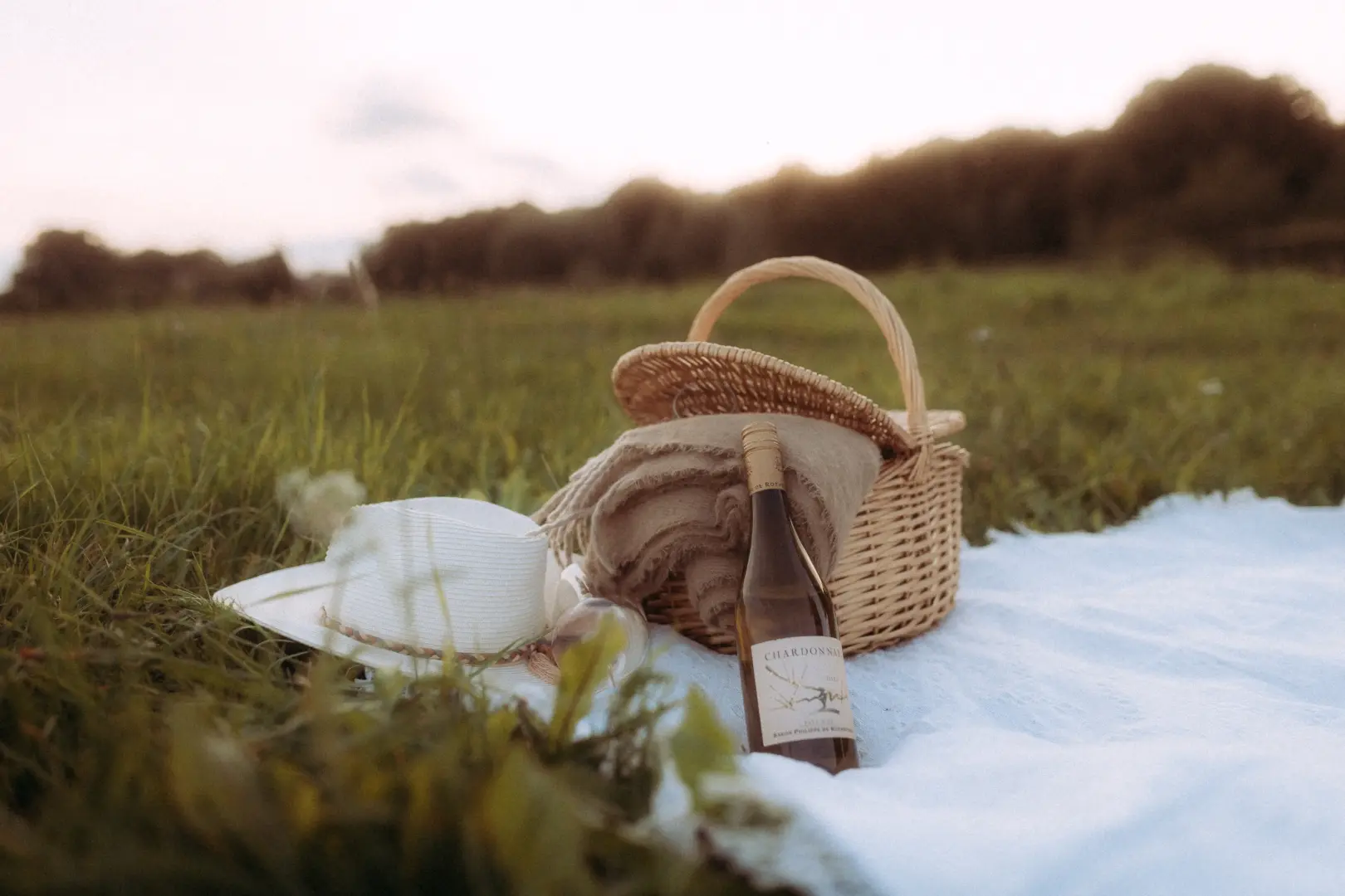 Ein idyllisches Picknick-Set auf einer Wiese bei Sonnenuntergang mit einem Strohhut, einer Decke, einem geflochtenen Korb und einer Flasche Chardonnay. Dieses stimmungsvolle Bild eignet sich perfekt als Inspiration für ein Paarshooting von einem Fotograf für Pärchen in Schwerin.