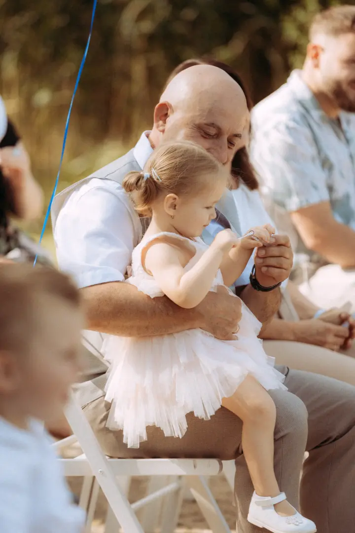 Ein glatzköpfiger Mann hält ein kleines Mädchen in einem zarten, hellrosa Tüllkleid auf dem Schoß, während sie gemeinsam einen Ehering betrachten. Das emotionale Familienfoto, eingefangen von einem Fotograf für Familien in Schwerin, zeigt eine liebevolle Szene bei einer Hochzeitsfeier.