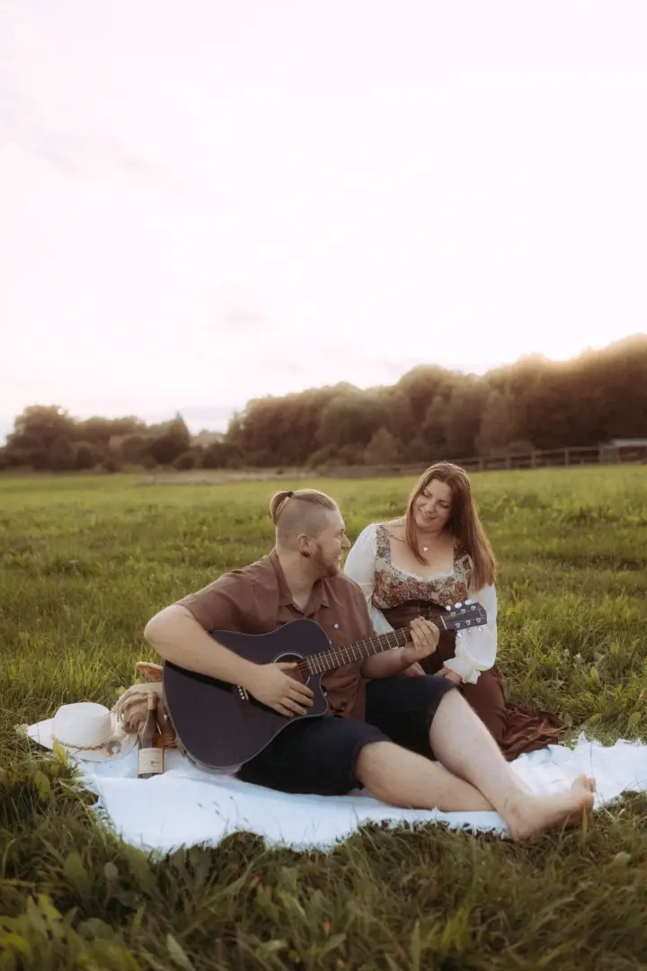 Ein Paar bei einem romantischen Picknick auf einer Wiese im goldenen Licht der Abendsonne. Der Mann spielt Gitarre, während die Frau ihn lächelnd ansieht. Eine stimmungsvolle Aufnahme für Pärchenfotos Schwerin, die eine intime und natürliche Verbindung zeigt.