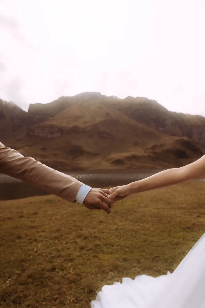 Ein Brautpaar hält sich vor einer dramatischen Bergkulisse an den Händen, wobei der Fokus auf der zärtlichen Geste liegt. Die Szene vermittelt eine intime Atmosphäre, perfekt eingefangen von einem kinematischen Hochzeitsfotografen in der Schweiz.