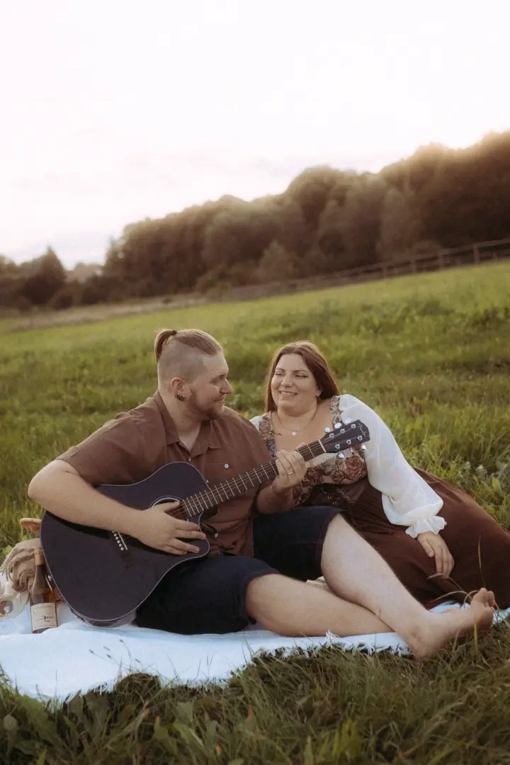 Ein glückliches Paar bei einem romantischen Picknick auf einer Wiese im goldenen Licht: Ein Mann spielt auf einer Akustikgitarre, während seine Partnerin ihn liebevoll anlächelt. Diese authentischen Pärchenfotos Schwerin fangen einen intimen, natürlichen Moment im Freien ein.