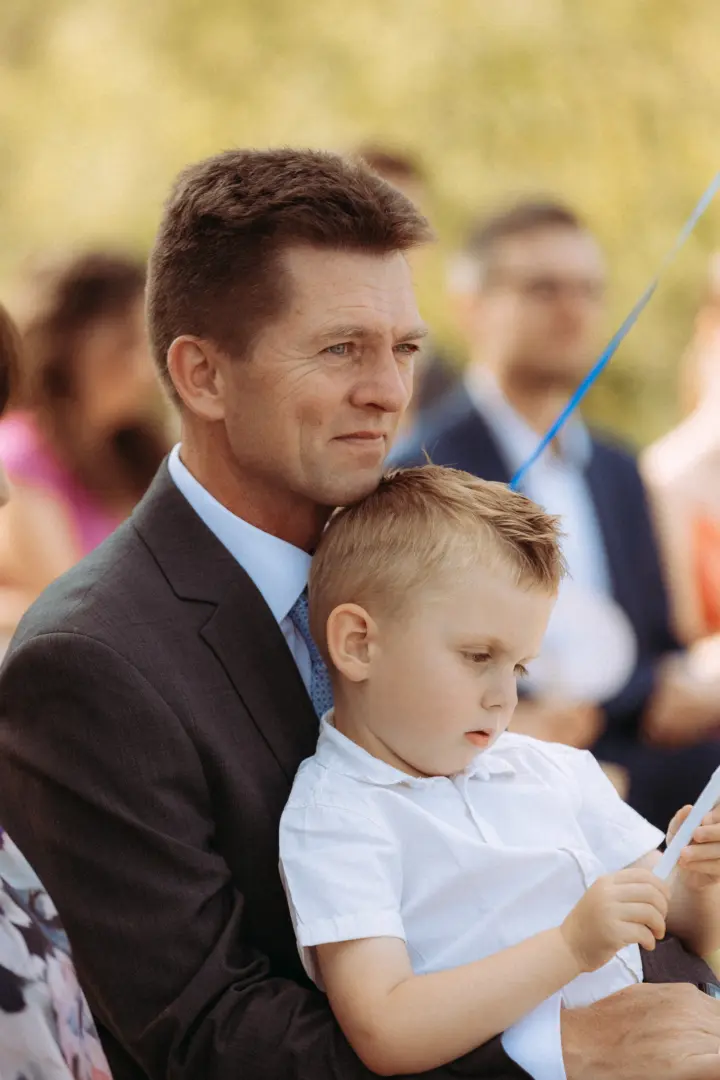 Ein liebevoller Familienmoment: Ein Vater im dunklen Anzug hält seinen kleinen Sohn in einem weißen Hemd im Arm, während beide gemeinsam bei einer Zeremonie zuschauen. Ein authentisches Familienfoto vom Fotograf für Familien in Schwerin.