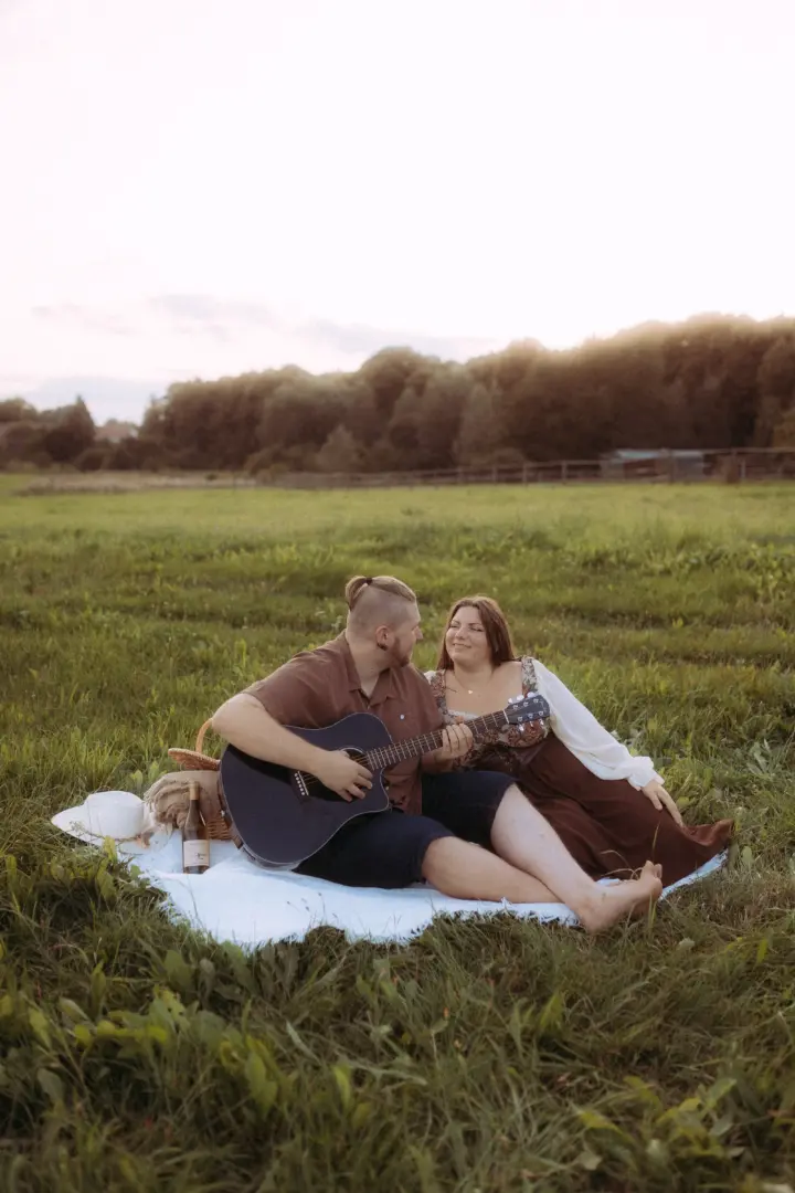 Ein Paar bei einem romantischen Picknick auf einer Wiese im goldenen Licht der Abendsonne. Der Mann spielt Gitarre, während die Frau ihn liebevoll ansieht. Ein stimmungsvolles Motiv für Pärchenfotos in Schwerin, das eine intime und natürliche Atmosphäre einfängt.