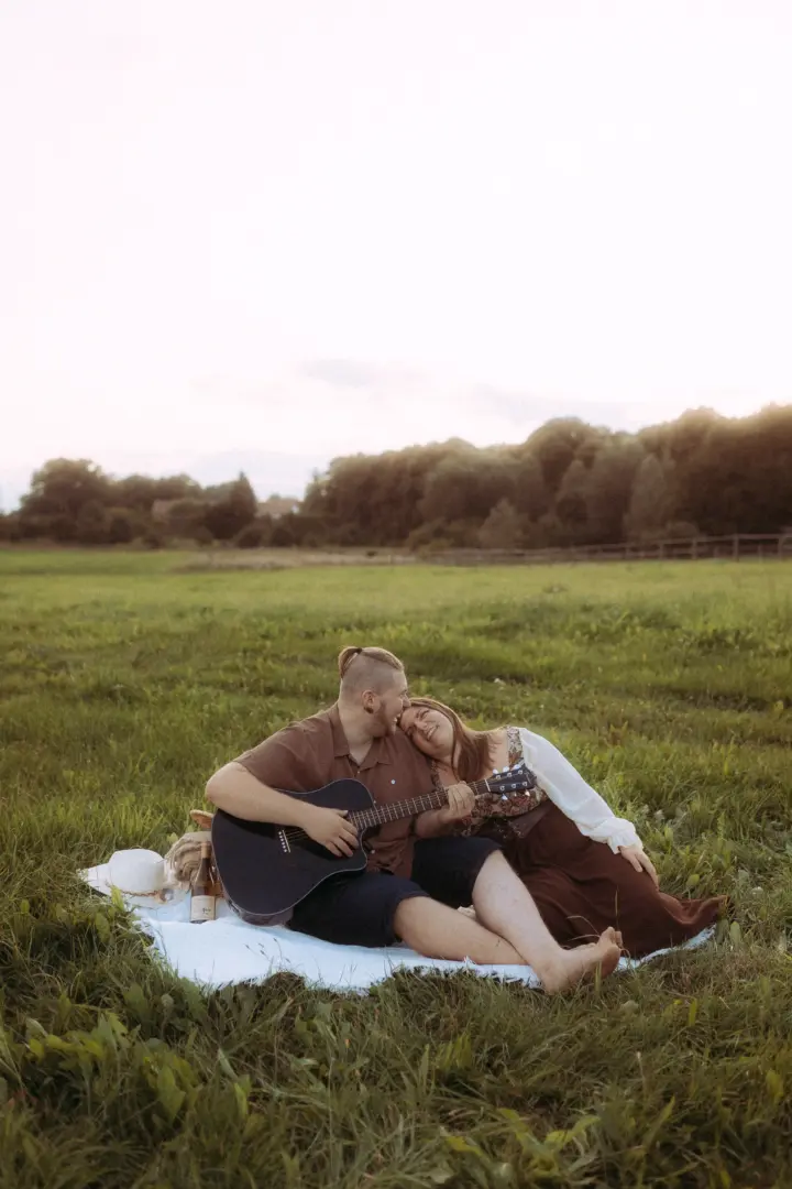 Ein glückliches Paar bei einem romantischen Picknick auf einer Wiese im goldenen Licht, wobei der Mann seiner Partnerin auf der Gitarre vorspielt. Diese stimmungsvollen Pärchenfotos Schwerin fangen die innige Verbindung und die entspannte Atmosphäre in der Natur perfekt ein.