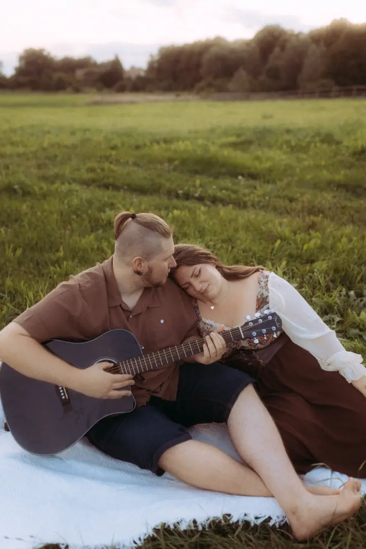 Ein Pärchen sitzt auf einer weißen Decke in einer grünen Wiese, während der Mann Gitarre spielt und die Frau ihren Kopf liebevoll an seine Schulter lehnt. Romantische Pärchenfotos Schwerin in natürlicher Atmosphäre bei Sonnenuntergang.