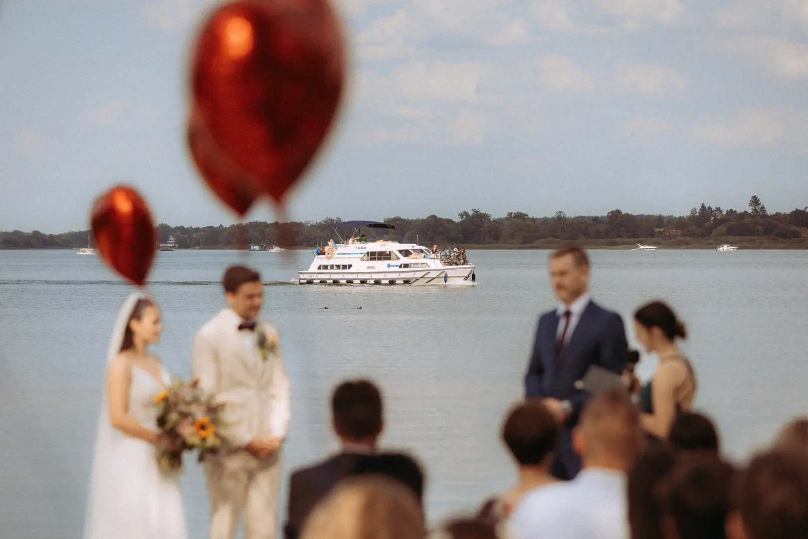 Ein Brautpaar steht während der Trauung am Ufer der Müritz, im Hintergrund zieht ein weißes Boot vorbei. Rote Luftballons im Vordergrund rahmen die Szene ein, während Gäste der Zeremonie zuschauen. Eine romantische Stimmung eingefangen von Ihrem Hochzeitsfotograf Müritz und MV.