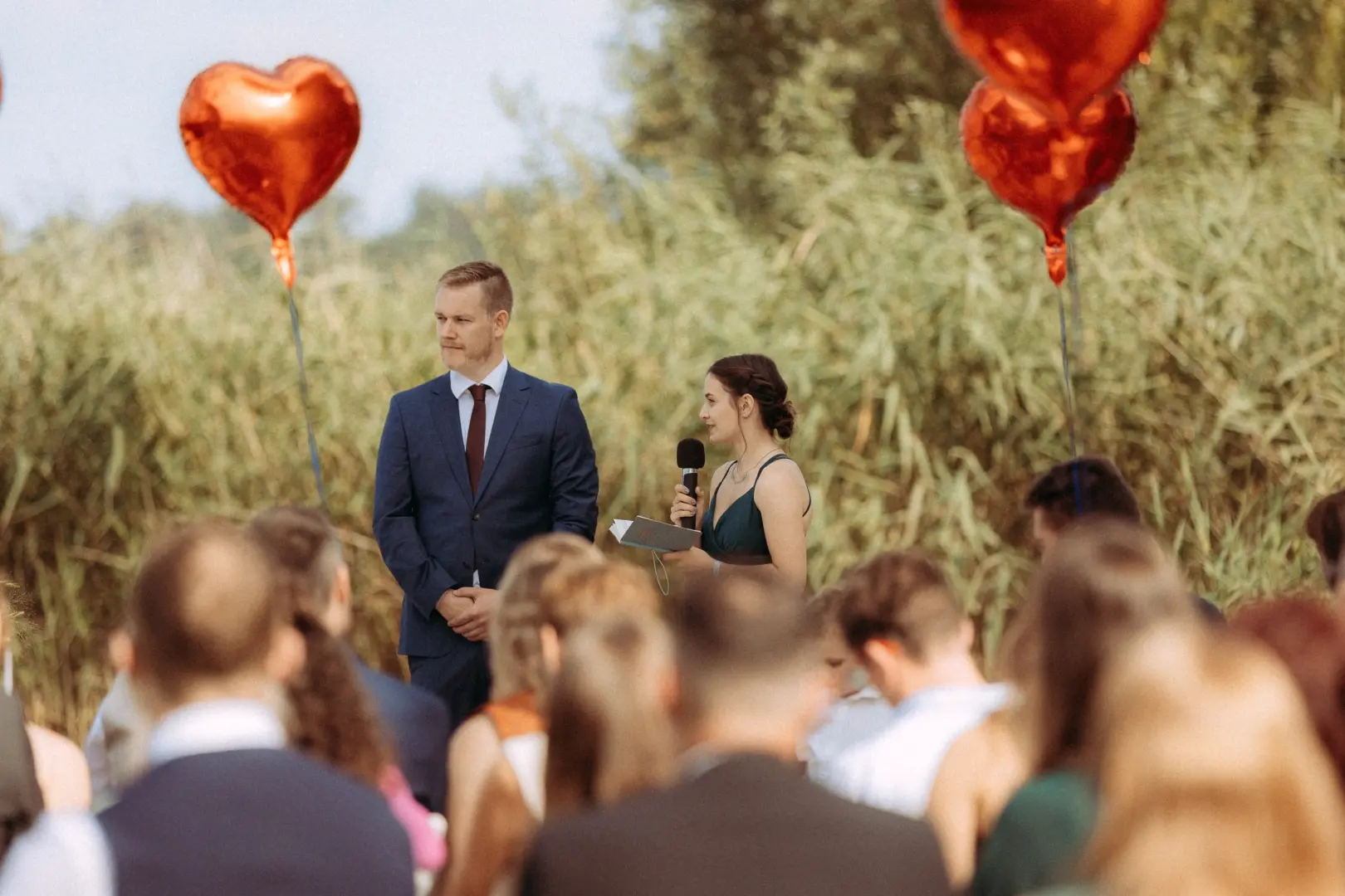 Eine Brautjungfer hält bei einer Hochzeitszeremonie im Freien eine Rede mit Mikrofon, während der Bräutigam im blauen Anzug daneben steht. Rote herzförmige Luftballons und hohes Schilfgras bilden die Kulisse für diesen Moment, festgehalten von einem Hochzeitsfotograf in Schwerin und MV.