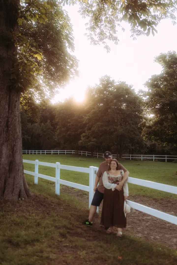 Ein glückliches Paar bei einem romantischen Fotoshooting an einem weißen Zaun auf einer grünen Wiese im warmen Licht der Abendsonne. Die beiden strahlen in einer natürlichen Umgebung, perfekt für authentische Pärchenfotos Schwerin.