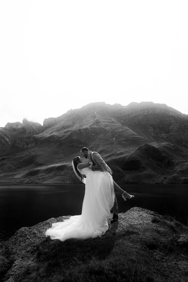 Ein Brautpaar in einer dramatischen Pose auf einem Felsen vor einem majestätischen Gebirgssee. Die Braut im weißen Kleid beugt sich zurück, während der Bräutigam sie hält. Kinematischer Hochzeitsfotograf in der Schweiz für unvergessliche Momente in atemberaubender Bergkulisse.