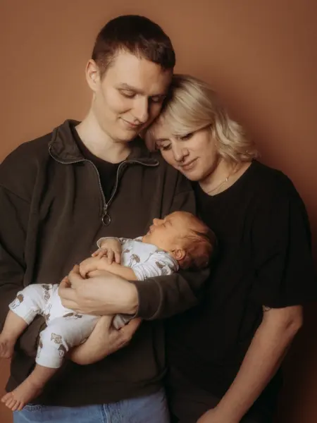 Ein junges Paar hält liebevoll sein neugeborenes Baby in einem Fotostudio in Schwerin. Die Eltern betrachten ihr Kind voller Zuneigung vor einem schlichten, braunen Hintergrund. Ein berührendes Familienfoto, das als Familienfotograf in Schwerin festgehalten wurde.