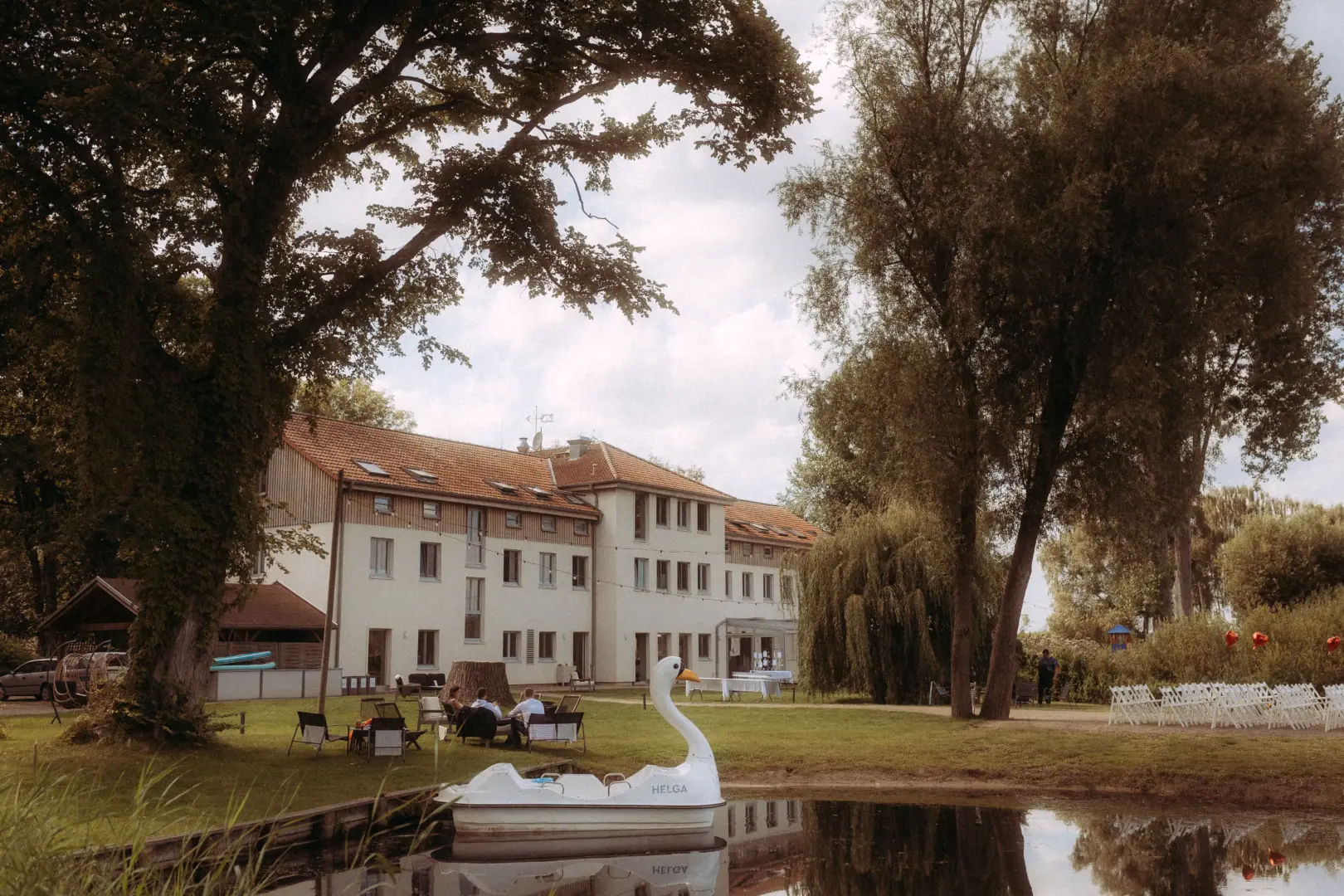 Ein weißes Tretboot in Schwanenform mit der Aufschrift „HELGA“ liegt in einem idyllischen Teich vor einem großen Landhaus. Im Hintergrund sind eine Hochzeitslocation mit Stuhlreihen auf der Wiese und Gäste zu sehen, eingefangen von einem Hochzeitsfotograf in Schwerin und MV.