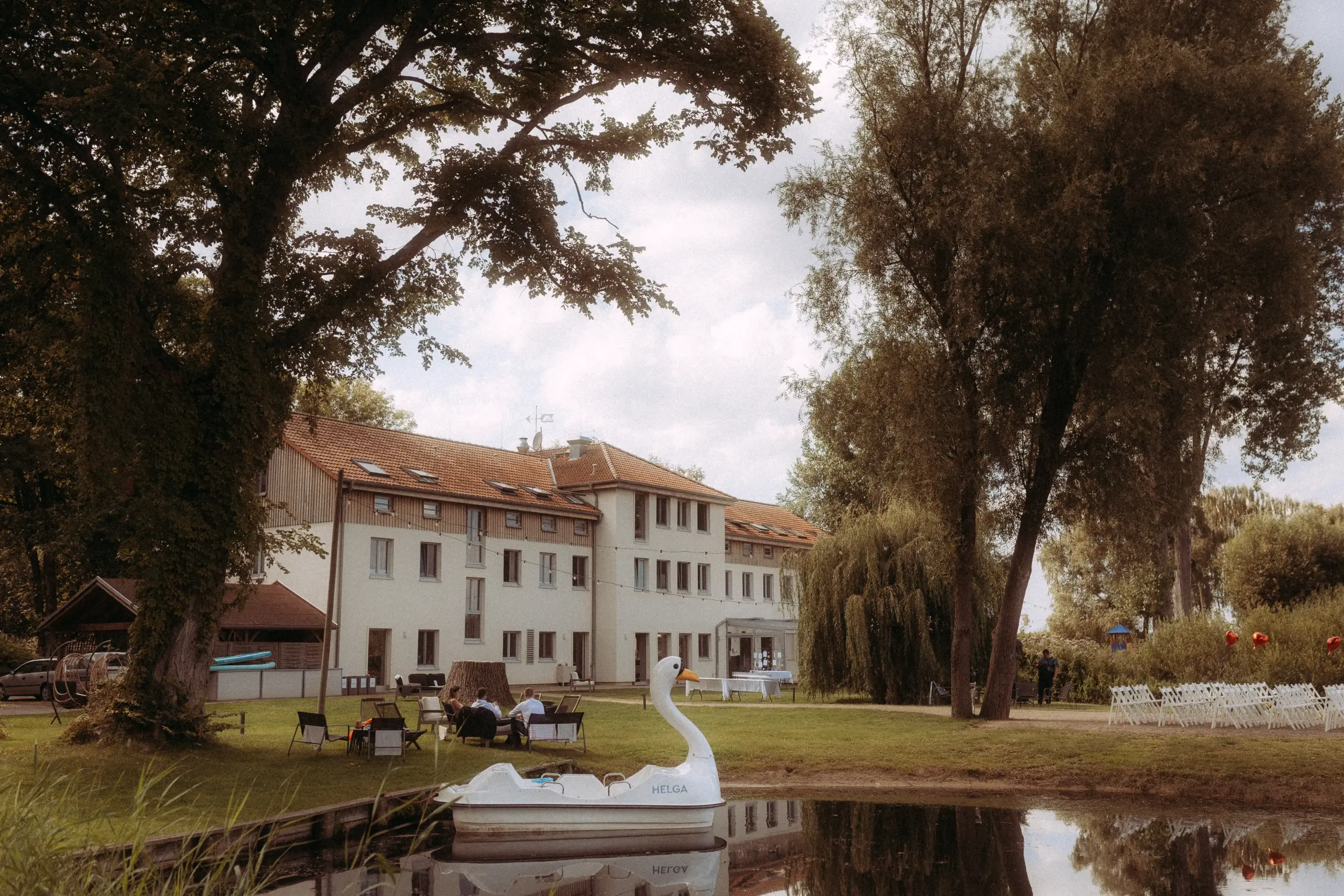 Ein weißes Tretboot in Schwanenform mit der Aufschrift „HELGA“ liegt in einem idyllischen Teich vor einem großen Landhaus. Im Hintergrund sind eine Hochzeitslocation mit Stuhlreihen auf der Wiese und Gäste zu sehen, eingefangen von einem Hochzeitsfotograf in Schwerin und MV. Ein weißes Tretboot in Schwanenform mit der Aufschrift „HELGA“ liegt in einem idyllischen Teich vor einem großen Landhaus. Im Hintergrund sind eine Hochzeitslocation mit Stuhlreihen auf der Wiese und Gäste zu sehen, eingefangen von einem Hochzeitsfotograf in Schwerin und MV.
