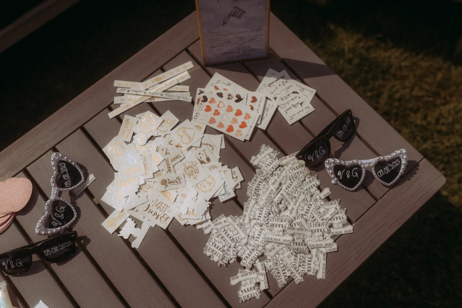 Ein Holztisch bei einer Hochzeit mit einer Auswahl an temporären Tattoos und herzförmigen Sonnenbrillen mit der Aufschrift „V&G 09.08.2025“. Diese liebevollen Details wurden von einem Hochzeitsfotograf in Schwerin und MV für die Gäste zur Unterhaltung bereitgestellt.