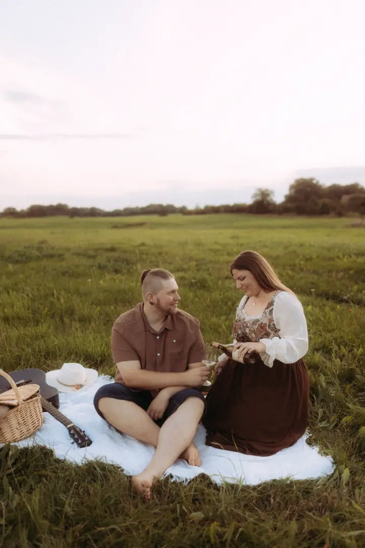 Ein Paar genießt ein romantisches Picknick auf einer grünen Wiese bei Sonnenuntergang. Die Frau schenkt Wein ein, während der Mann sie liebevoll ansieht. Neben ihnen liegen eine Gitarre und ein Picknickkorb. Authentische Pärchenfotos Schwerin für unvergessliche Momente im Freien.