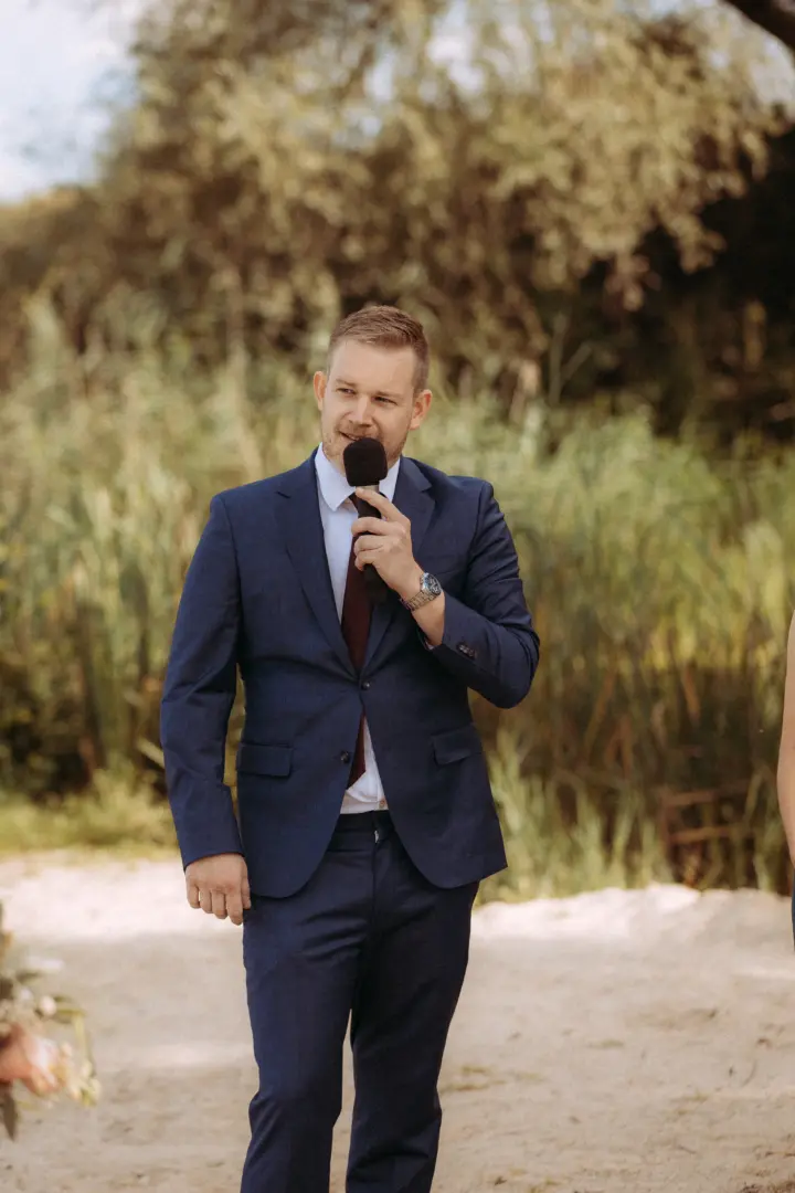 Ein eleganter Redner im dunkelblauen Anzug mit Krawatte hält ein Mikrofon und spricht während einer Outdoor-Hochzeitszeremonie. Im Hintergrund ist eine natürliche Kulisse aus Schilf und Bäumen zu sehen. Hochzeitsfotograf in Schwerin und MV.