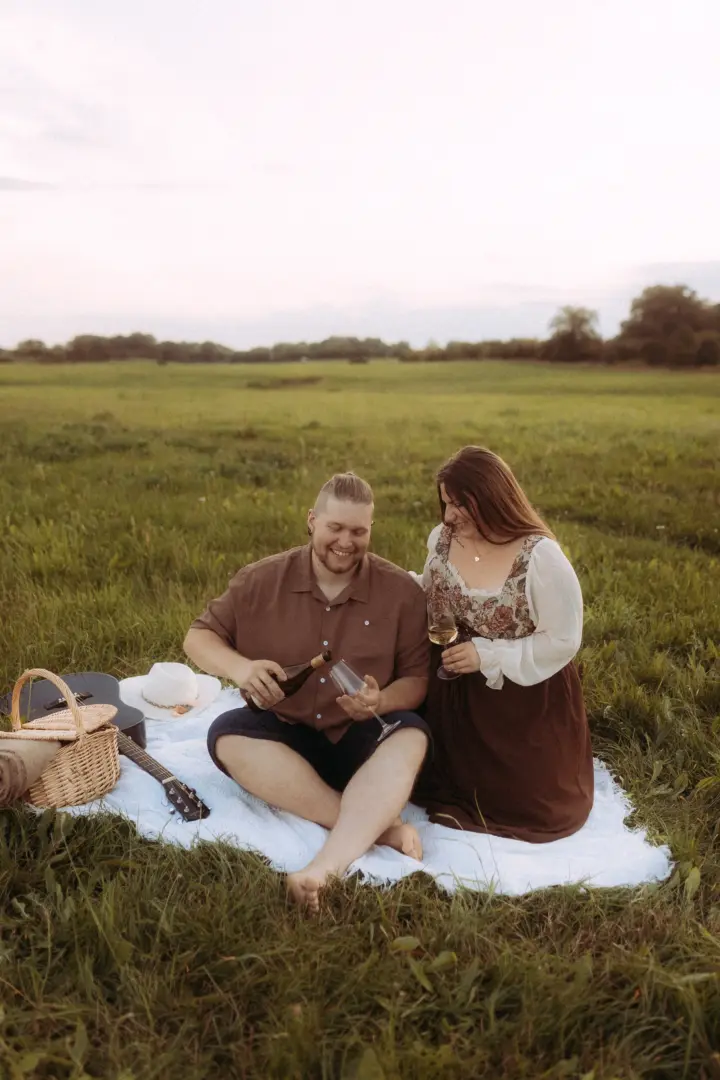 Ein glückliches Paar bei einem romantischen Picknick auf einer grünen Wiese bei Sonnenuntergang. Der Mann schenkt Wein in ein Glas ein, während die Frau lächelnd zusieht. Eine Gitarre und ein Korb liegen auf der Decke. Authentische Pärchenfotos Schwerin für unvergessliche Momente.