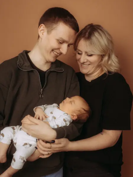 Ein junges Paar hält liebevoll sein schlafendes Neugeborenes im Arm, während beide glücklich auf das Baby blicken. Das professionelle Familienfoto entstand in einem Fotostudio in Schwerin vor einem warmen, braunen Hintergrund.