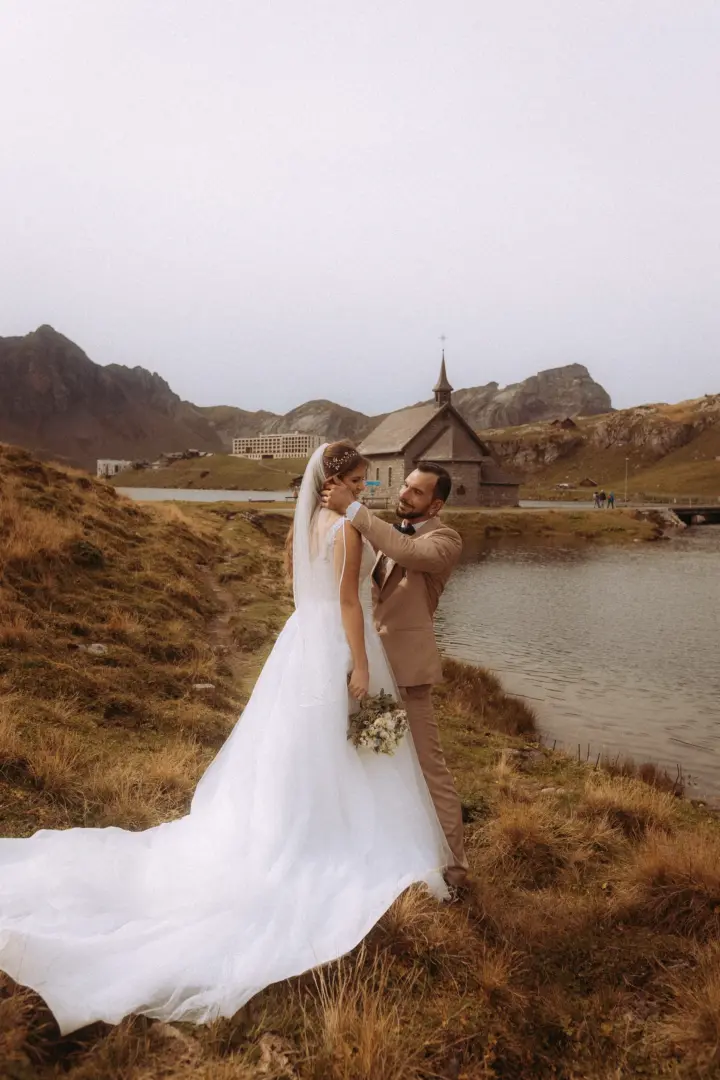 Ein Brautpaar in romantischer Pose vor einer Kapelle am Bergsee. Der Bräutigam hält liebevoll das Gesicht der Braut, die ein weißes Brautkleid trägt. Diese stimmungsvolle Aufnahme entstand durch einen kinematischen Hochzeitsfotograf in der Schweiz in einer atemberaubenden Berglandschaft.