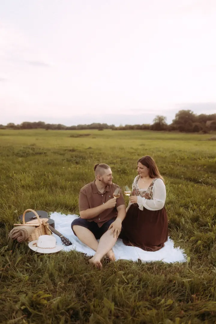 Ein Paar genießt ein romantisches Picknick auf einer weiten Wiese bei Sonnenuntergang. Sie stoßen mit Weingläsern an, während eine Gitarre und ein Picknickkorb neben ihnen auf einer Decke liegen. Authentische Pärchenfotos Schwerin, die eine intime und entspannte Stimmung einfangen.