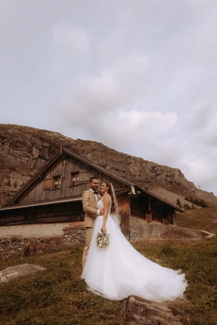 Ein glückliches Brautpaar steht vor einer rustikalen Holzhütte in den Alpen, während die Braut ein weißes Brautkleid trägt und der Bräutigam einen beigen Anzug. Diese Aufnahme zeigt die Arbeit eines kinematischen Hochzeitsfotografen in der Schweiz.