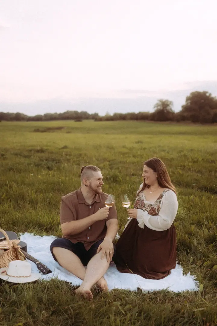 Ein glückliches Paar stößt bei einem romantischen Picknick auf einer weiten Wiese mit Weingläsern an. Sie tragen herbstliche Kleidung, neben ihnen liegen ein Picknickkorb und eine Gitarre. Authentische Pärchenfotos Schwerin, eingefangen in einer idyllischen Naturkulisse.