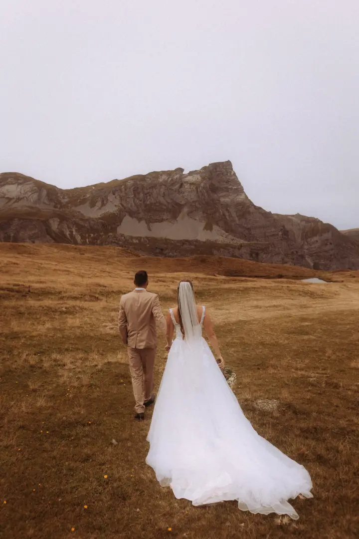 Ein Brautpaar geht Hand in Hand durch eine weite, herbstliche Berglandschaft in Richtung eines majestätischen Gipfels. Die Braut trägt ein weißes Hochzeitskleid mit Schleier, der Bräutigam einen beigen Anzug. Kinematischer Hochzeitsfotograf in der Schweiz.