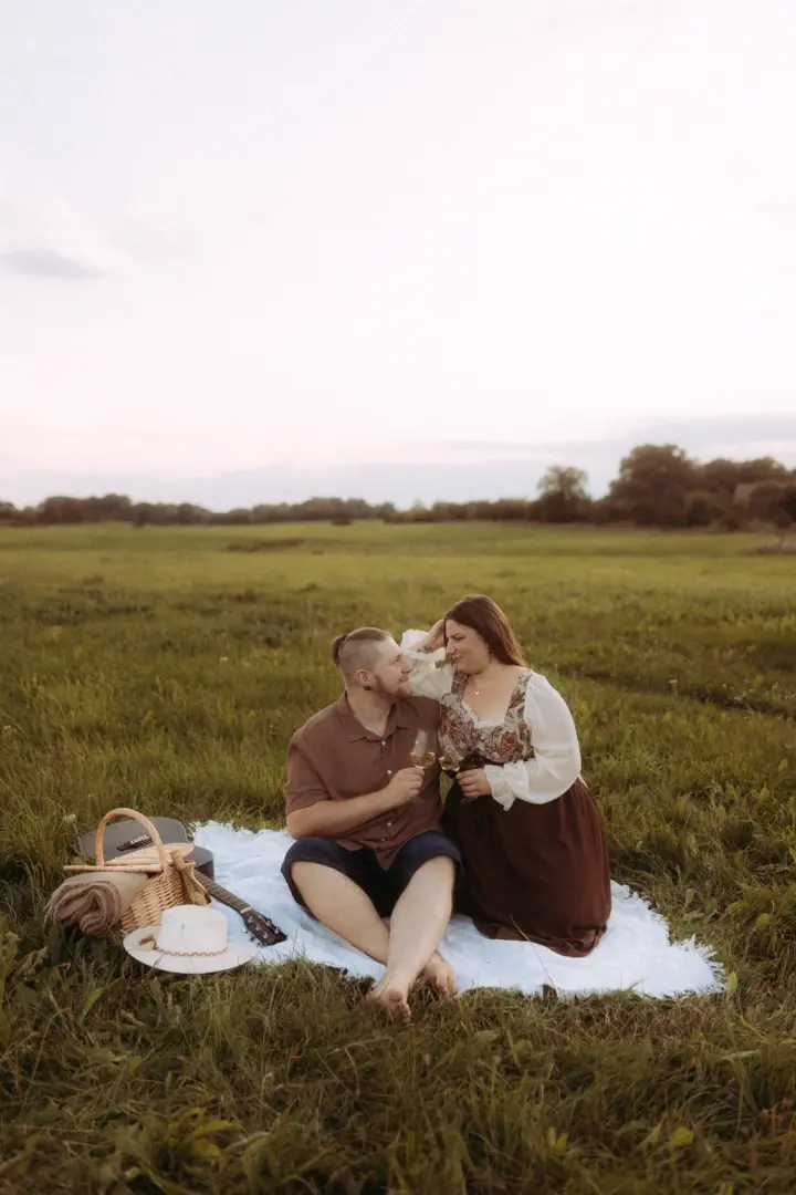 Ein Paar genießt ein romantisches Picknick auf einer grünen Wiese bei Sonnenuntergang. Sie stoßen mit Weingläsern an, neben ihnen stehen ein Picknickkorb und eine Gitarre. Authentische Pärchenfotos Schwerin, die eine liebevolle Stimmung in der Natur einfangen.