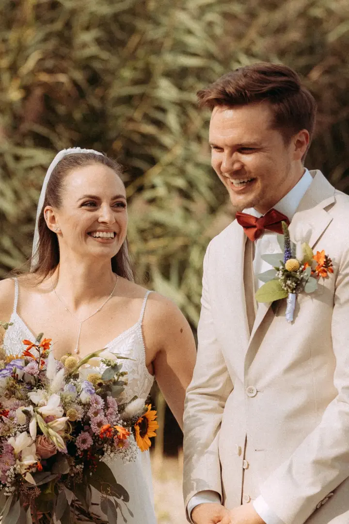 Ein glückliches Brautpaar bei der Trauung im Freien: Die Braut trägt ein weißes Brautkleid mit Blumenstrauß, der Bräutigam einen beigen Anzug mit roter Fliege. Ein emotionaler Moment festgehalten von Ihrem Hochzeitsfotograf in Schwerin und MV.