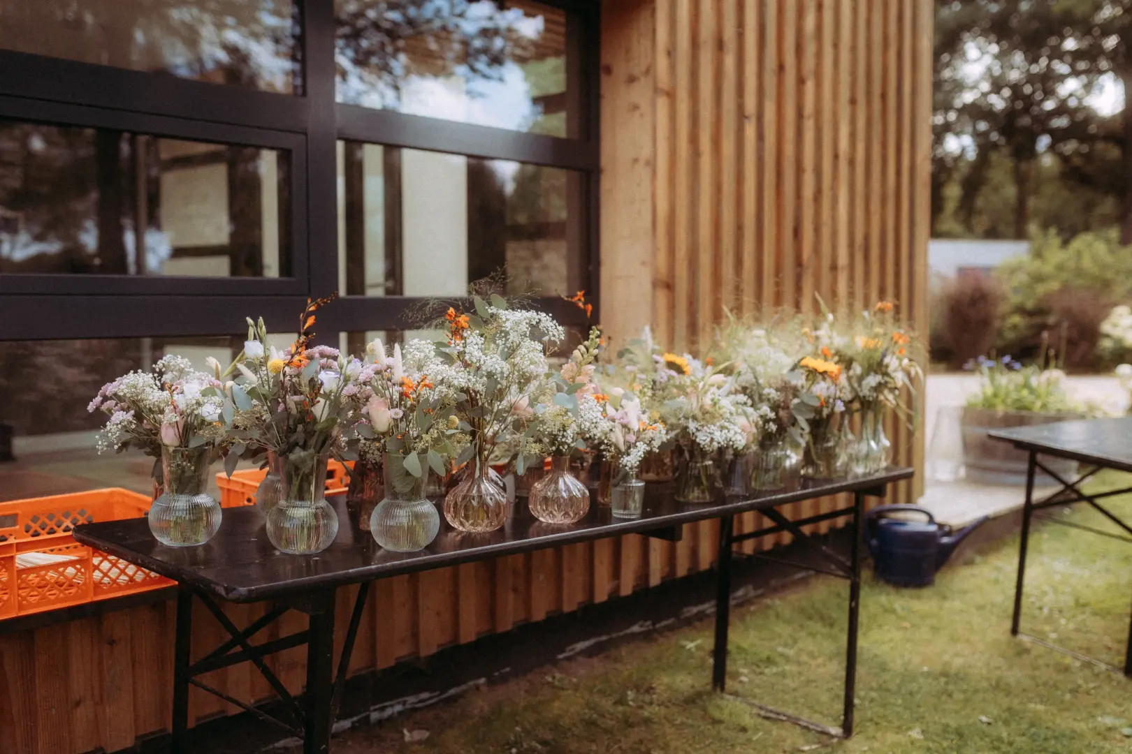 Eine Reihe von kleinen, mit bunten Wildblumen gefüllten Glasvasen steht auf einem schwarzen Tisch vor einer modernen Holzfassade. Die florale Dekoration sorgt für eine natürliche Hochzeitsstimmung, festgehalten von einem Hochzeitsfotograf in Schwerin und MV.
