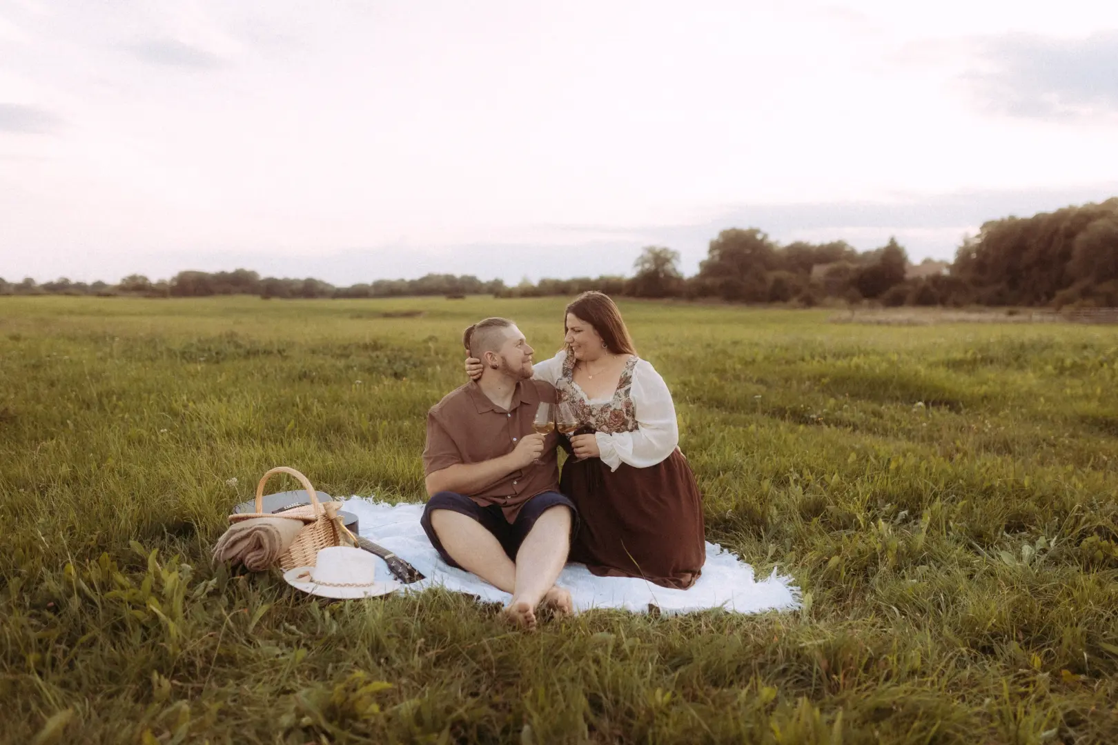 Ein glückliches Paar bei einem romantischen Picknick auf einer weitläufigen Wiese während des Sonnenuntergangs. Die beiden halten Weingläser in der Hand und genießen gemeinsame Zeit. Stimmungsvolle Pärchenfotos Schwerin für unvergessliche Momente im Freien.